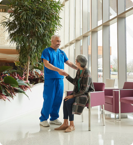 A male clinician in scrubs greeting an older female in a hospital waiting room.
